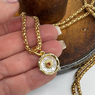 Gold necklace with a sun and heart pendant held by a hand on a wooden surface