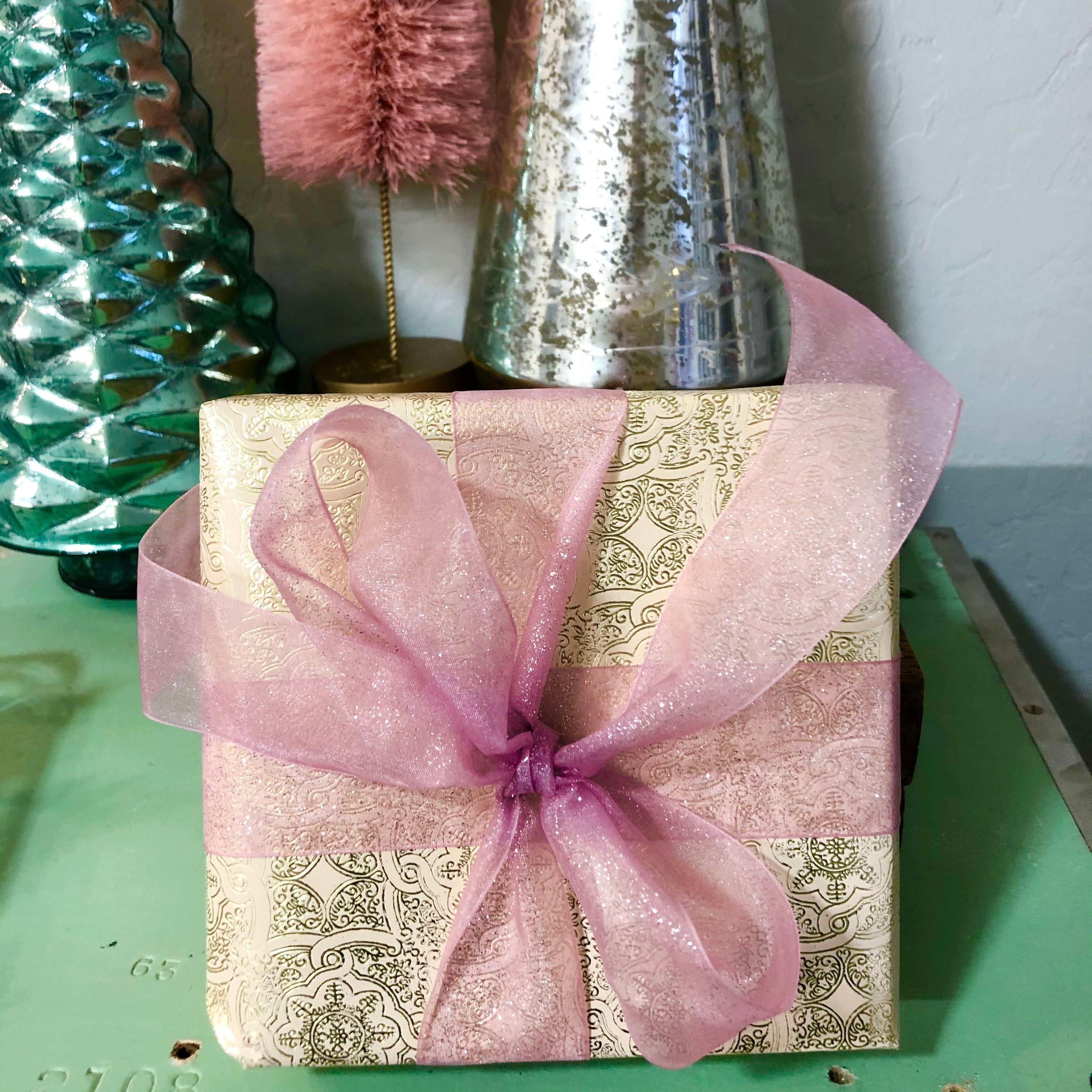 A gift box wrapped in gold and blue patterned wrapping paper with a pink bow on top. The box is placed on a green surface, and there are decorative items in the background.