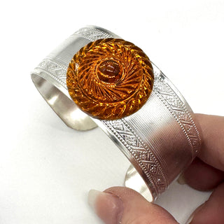 Silver bracelet with an orange lucite stone with a circular design held by a hand on a white background