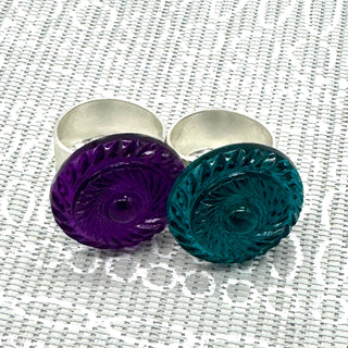 Two lucite cocktail rings, one purple and one teal, on a textured white background.