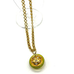 A gold chain necklace with a round pendant featuring a lime green outer ring and a gold-toned center decorated with small clear rhinestones, displayed on a clear stand against a white background.