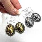A close-up of a hand holding a white card displaying two pairs of oval stud earrings on a white background. The pairs are gold-toned and silver-toned with ornate beaded borders and smooth oval centers.