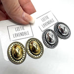 A close-up of a hand holding a white card printed with the brand name 'LEETIE LOVENDALE' above a pair of oval stud earrings. The card also shows a second pair of matching earrings.