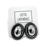 A pair of oval stud earrings with shiny silver-toned centers and dark textured outer borders, displayed on a white branded backing card that reads LEETIE LOVENDALE.