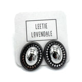 A pair of oval stud earrings with shiny silver-toned centers and dark textured outer borders, displayed on a white branded backing card that reads LEETIE LOVENDALE.