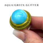 Aqua and green glittery ring held by a hand against a white background