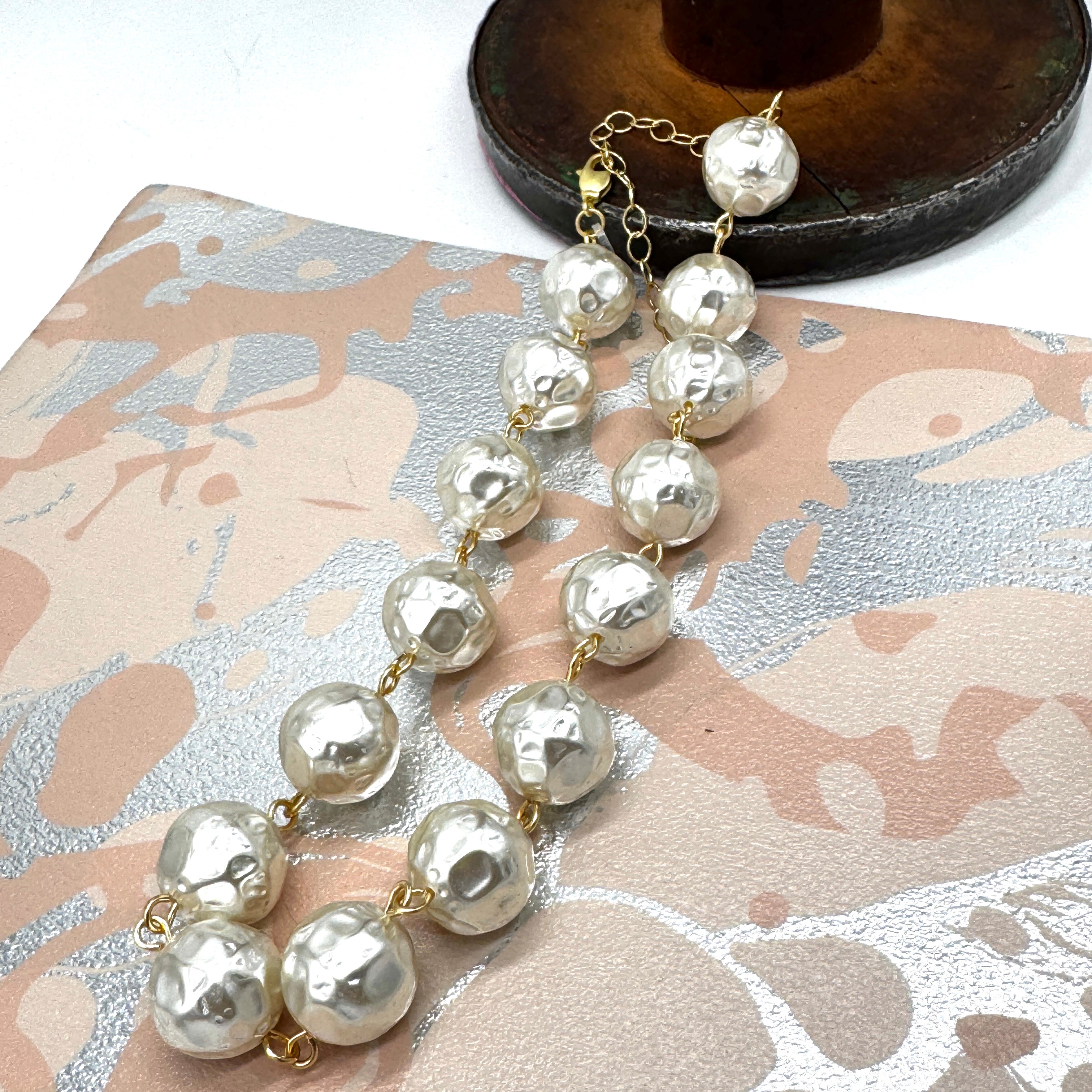 A strand of irregular white resin pearls with textured, hammered finishes, connected by gold-tone chains and clasps, displayed on a patterned beige and silver surface.