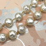 A close-up of a pearl-like beaded chain with hammered, textured round beads connected by small gold-tone links, laid diagonally across a patterned surface with pink, beige, and silver tones.