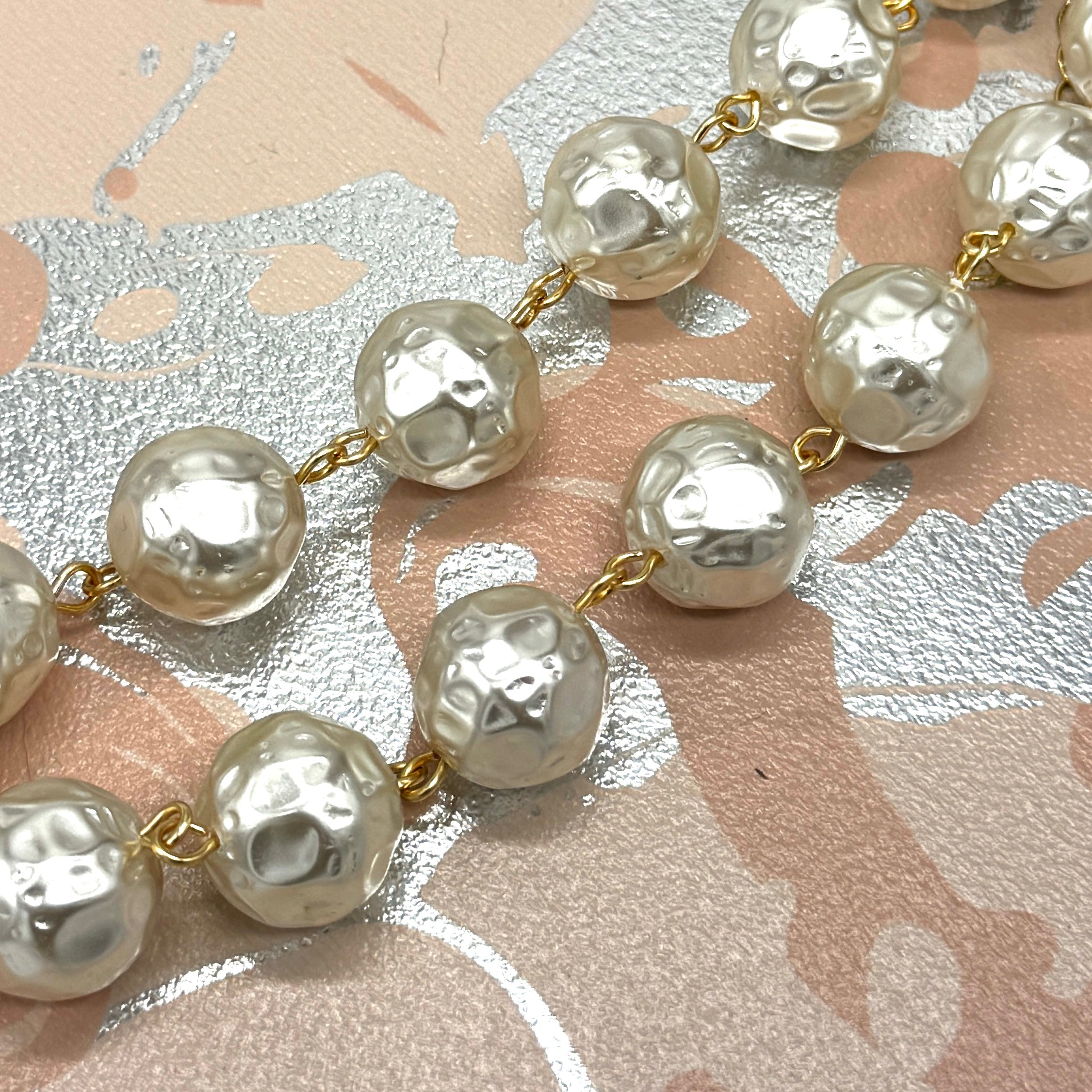 A close-up of a pearl-like beaded chain with hammered, textured round beads connected by small gold-tone links, laid diagonally across a patterned surface with pink, beige, and silver tones.
