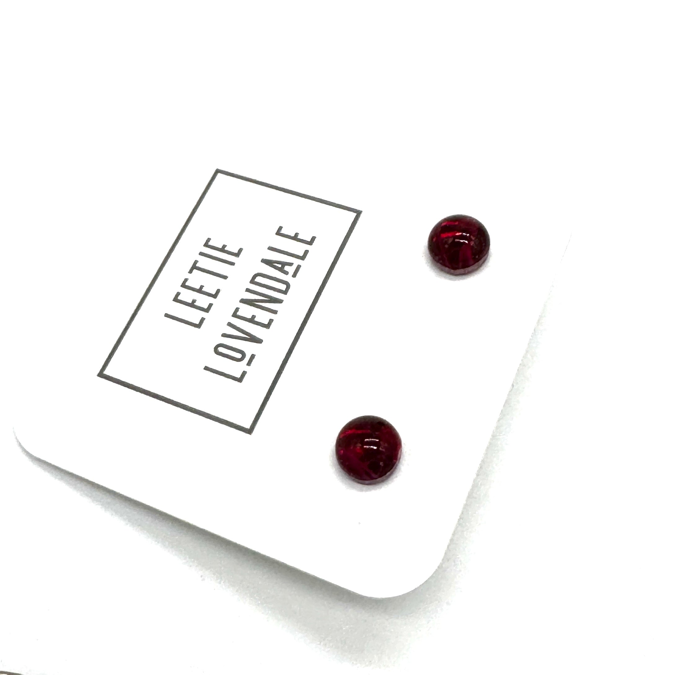 A pair of small round stud earrings in deep red resin, displayed on a white branded card that reads 'LEETIE LOVENDALE'.