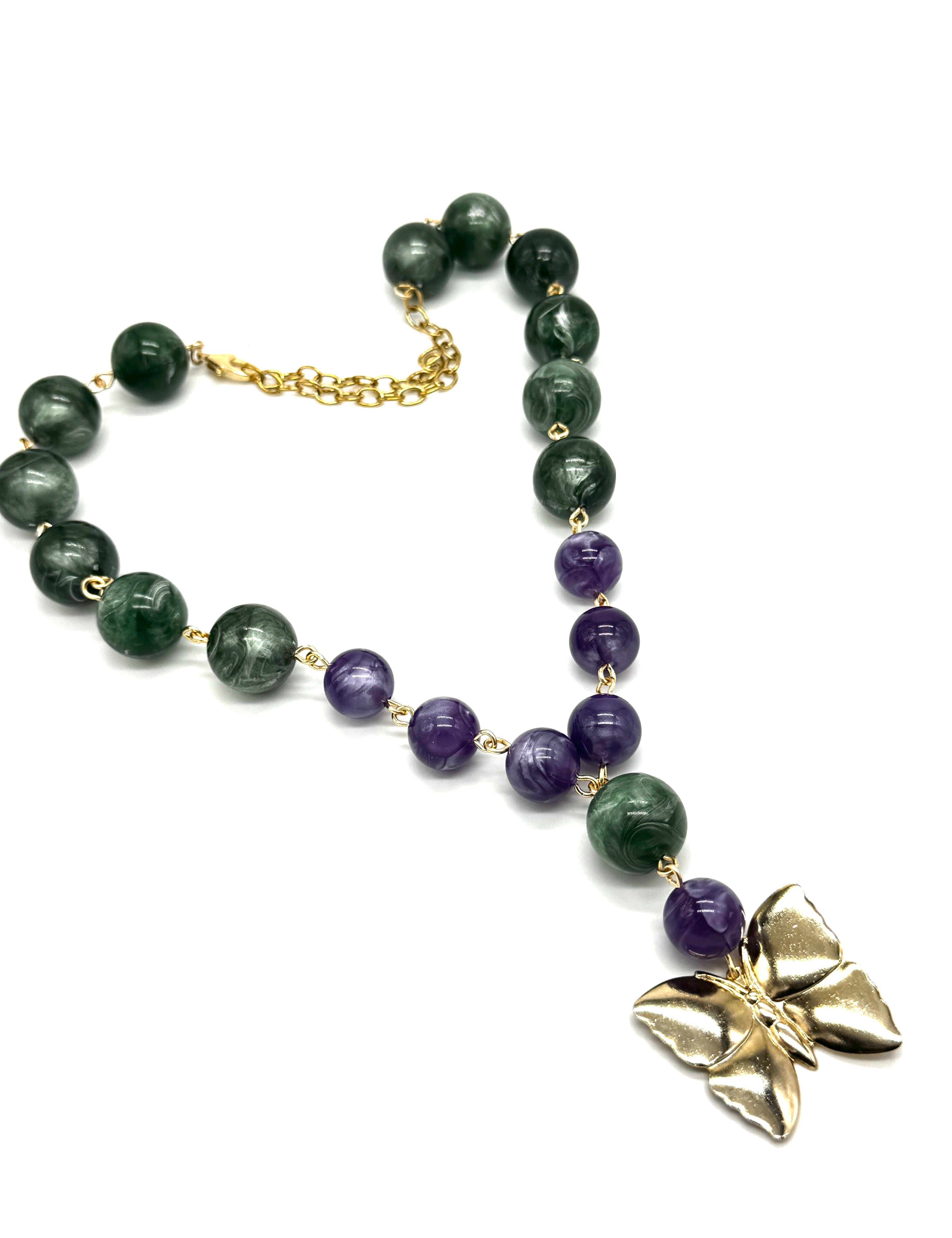 A beaded necklace featuring dark green and purple lucite beads connected by small gold links, with a gold butterfly pendant and gold chain clasp, shown on a plain white background.