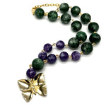 A beaded necklace featuring dark green lucite beads, purple amethyst beads, and a textured gold butterfly pendant, displayed on a clean white background.