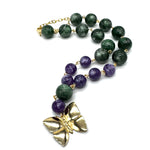 A beaded necklace featuring dark green and purple lucite beads separated by small gold links, with a large textured gold butterfly pendant at the end, displayed on a clean white background.