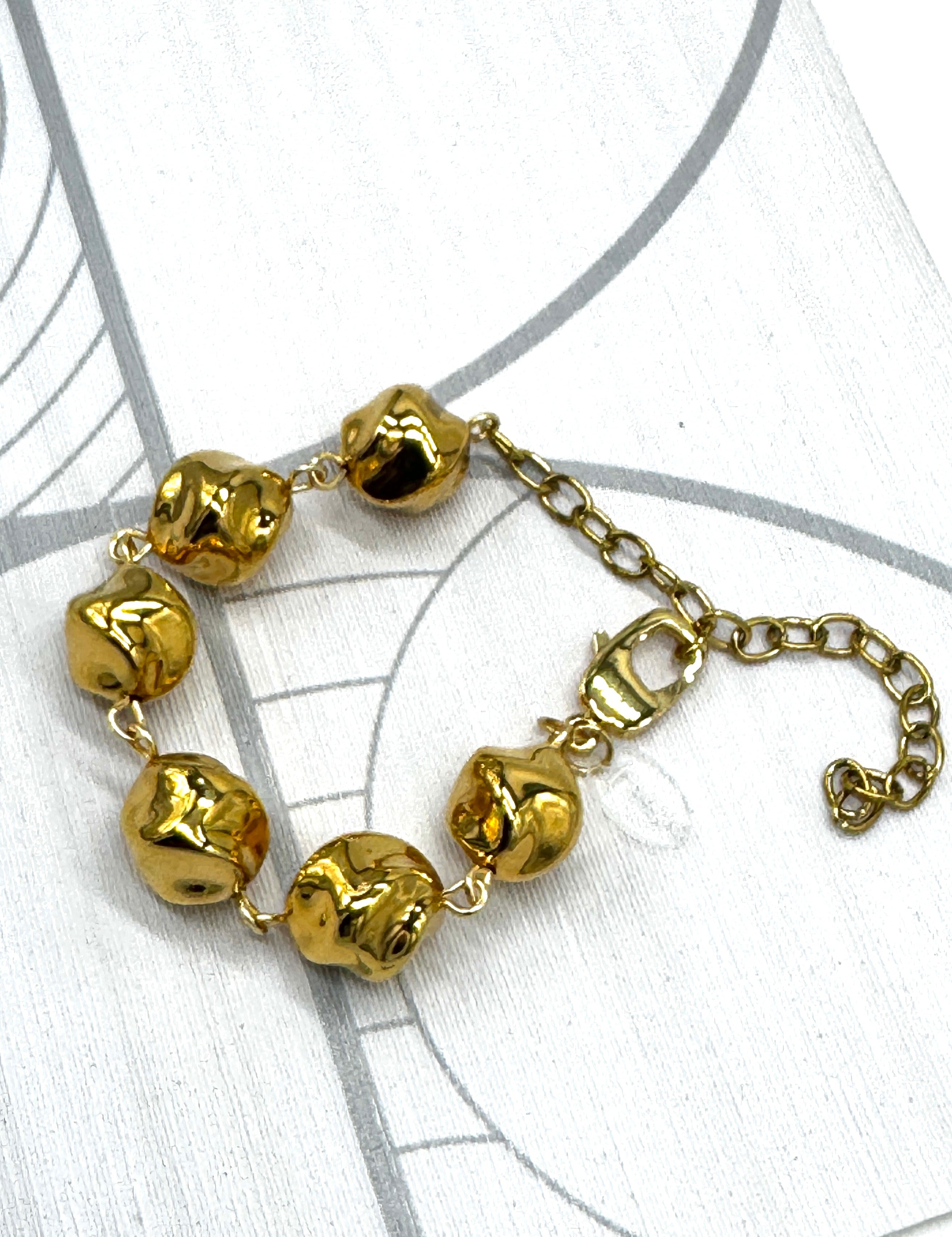 A gold-toned beaded bracelet with six irregular, polished beads connected by a delicate chain and extender clasp, displayed on a white background with faint gray circular line patterns.
