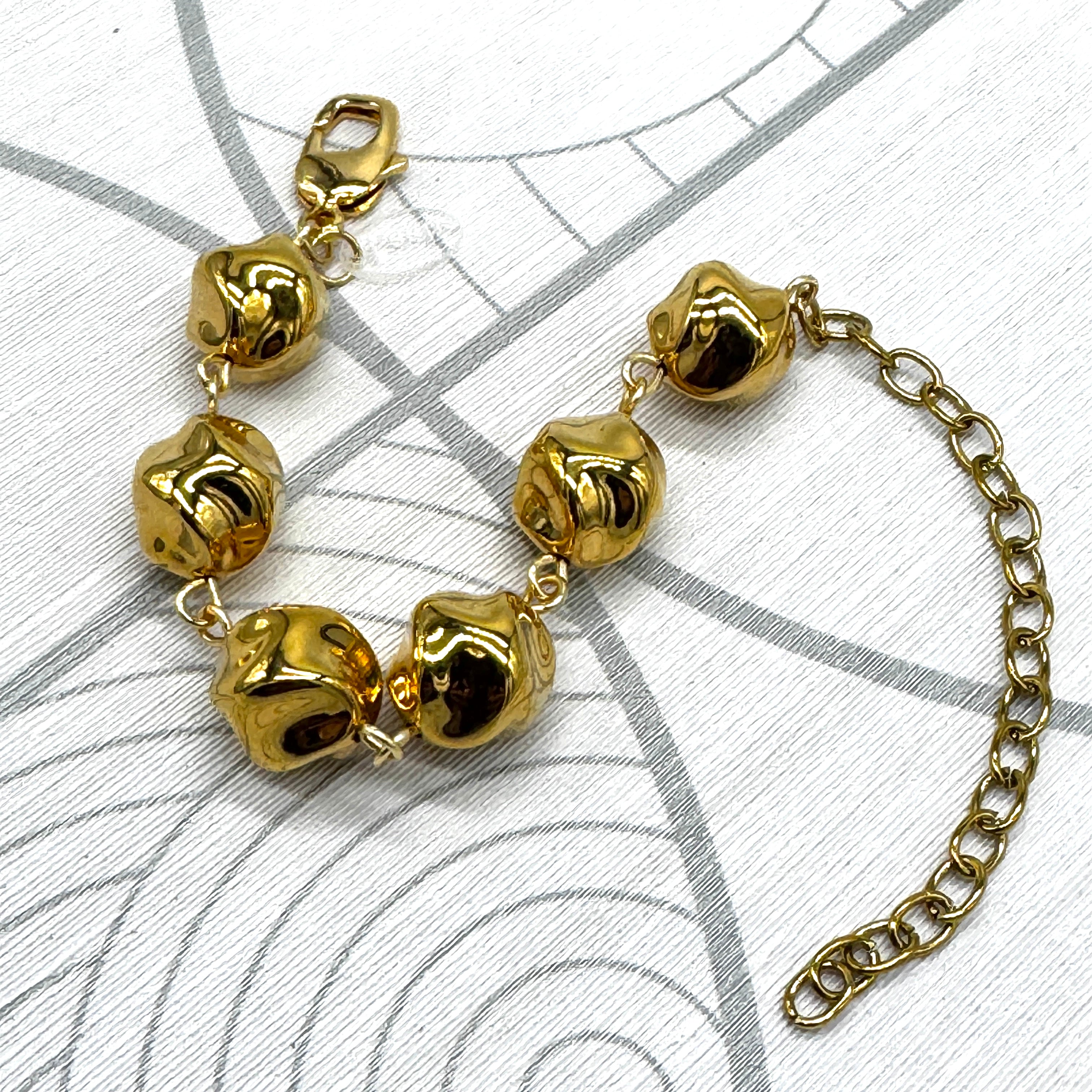 A gold-tone metal bracelet featuring textured, polished twisted round-shaped beads connected by small links, with a lobster clasp on one end and an adjustable extension chain on the other, displayed on a white patterned surface.